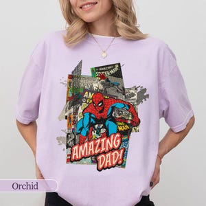 May include: Orchid-colored t-shirt featuring a comic book-style graphic of Spider-Man with the text "AMAZING DAD!". The design incorporates red, blue, and black colors. The shirt is a casual wear item.