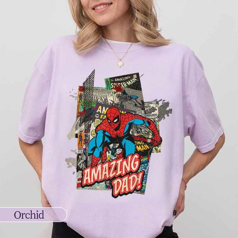 May include: Orchid-colored t-shirt featuring a comic book-style graphic of Spider-Man with the text "AMAZING DAD!". The design incorporates red, blue, and black colors. The shirt is a casual wear item.