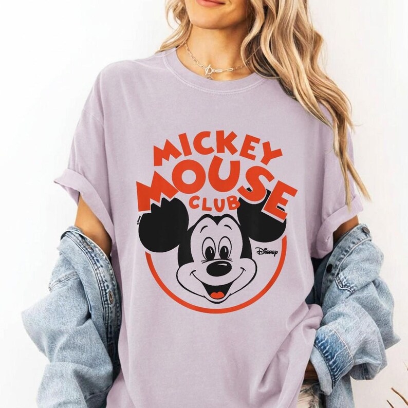 May include: A light purple t-shirt featuring the Mickey Mouse Club logo in red and black. The design includes Mickey Mouse's face inside a red circle, with the Disney logo below. The shirt has rolled-up sleeves.