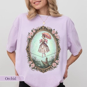 May include: Orchid-colored t-shirt featuring a framed illustration of a woman with an umbrella, standing on a tightrope above a crocodile. The frame is adorned with roses. The word "Orchid" is printed at the bottom.