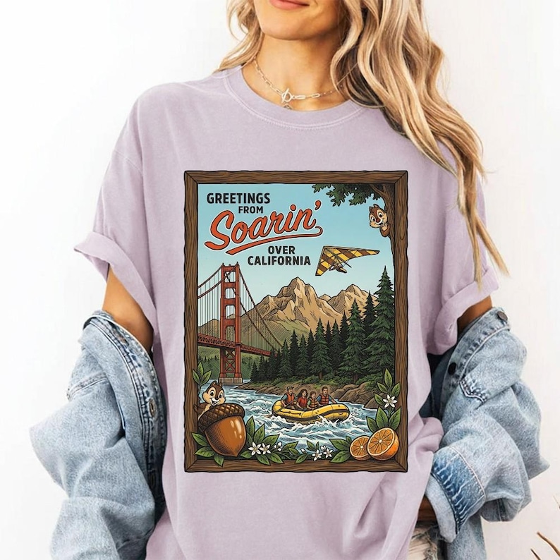 Puede incluir: Camiseta lila claro con un dise&ntilde;o enmarcado. El dise&ntilde;o dice "GREETINGS FROM Soarin' OVER CALIFORNIA" y muestra el Golden Gate Bridge, monta&ntilde;as, un ala delta y un r&iacute;o con una balsa.