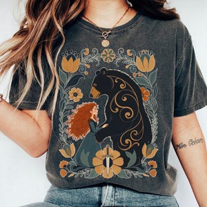 Op de afbeelding: Donkergrijs t-shirt met een bloemenontwerp met een beer en een vrouw met rood haar. De beer is zwart met gouden accenten en de vrouw draagt een blauwe jurk. Het ontwerp bevat gele en groene bloemelementen.
