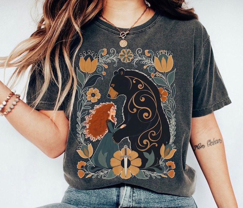 Op de afbeelding: Donkergrijs t-shirt met een bloemenontwerp met een beer en een vrouw met rood haar. De beer is zwart met gouden accenten en de vrouw draagt een blauwe jurk. Het ontwerp bevat gele en groene bloemelementen.