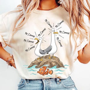 May include: Cream-colored t-shirt featuring a cartoon illustration of two seagulls on a rock, with the text "Mr. Connor" emanating from them. Below, an orange and white fish swims in blue water. The shirt is a casual wear item.