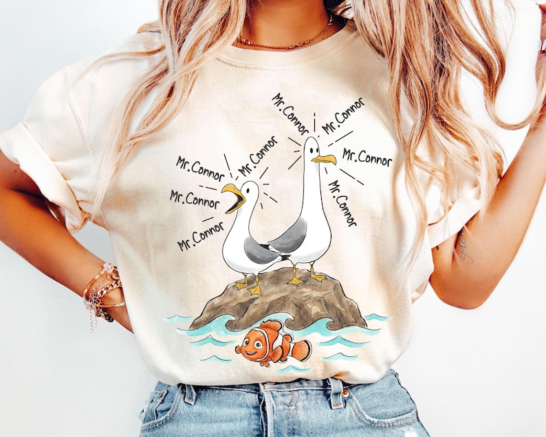 May include: Cream-colored t-shirt featuring a cartoon illustration of two seagulls on a rock, with the text "Mr. Connor" emanating from them. Below, an orange and white fish swims in blue water. The shirt is a casual wear item.