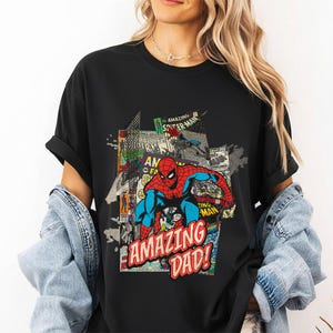 May include: Black t-shirt featuring a colorful comic book style graphic of Spider-Man with the text "AMAZING DAD!". The design includes comic book panels and the words "AMAZING SPIDER-MAN". The shirt has short sleeves and a crew neck.