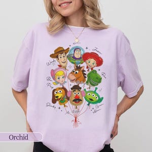 Puede incluir: Camiseta color orqu&iacute;dea con un gr&aacute;fico de personajes de Toy Story como globos. El dise&ntilde;o incluye a Woody, Buzz Lightyear, Jessie, Rex y otros, con sus nombres escritos debajo. La palabra "Orchid" est&aacute; impresa en la parte inferior.