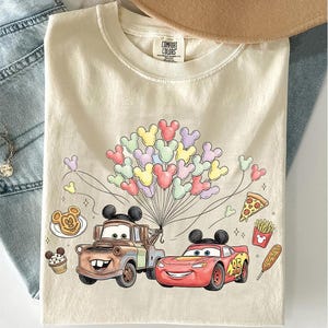 May include: Cream-coloured t-shirt with a graphic of two cartoon cars, Mater and Lightning McQueen, and Mickey Mouse balloons. The shirt also has food illustrations like pizza and fries. The tag reads "Comfort Colors".