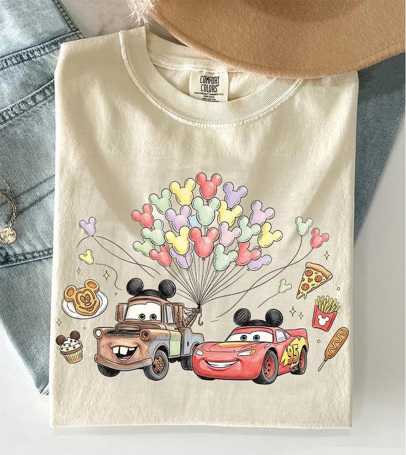 May include: Cream-coloured t-shirt with a graphic of two cartoon cars, Mater and Lightning McQueen, and Mickey Mouse balloons. The shirt also has food illustrations like pizza and fries. The tag reads "Comfort Colors".