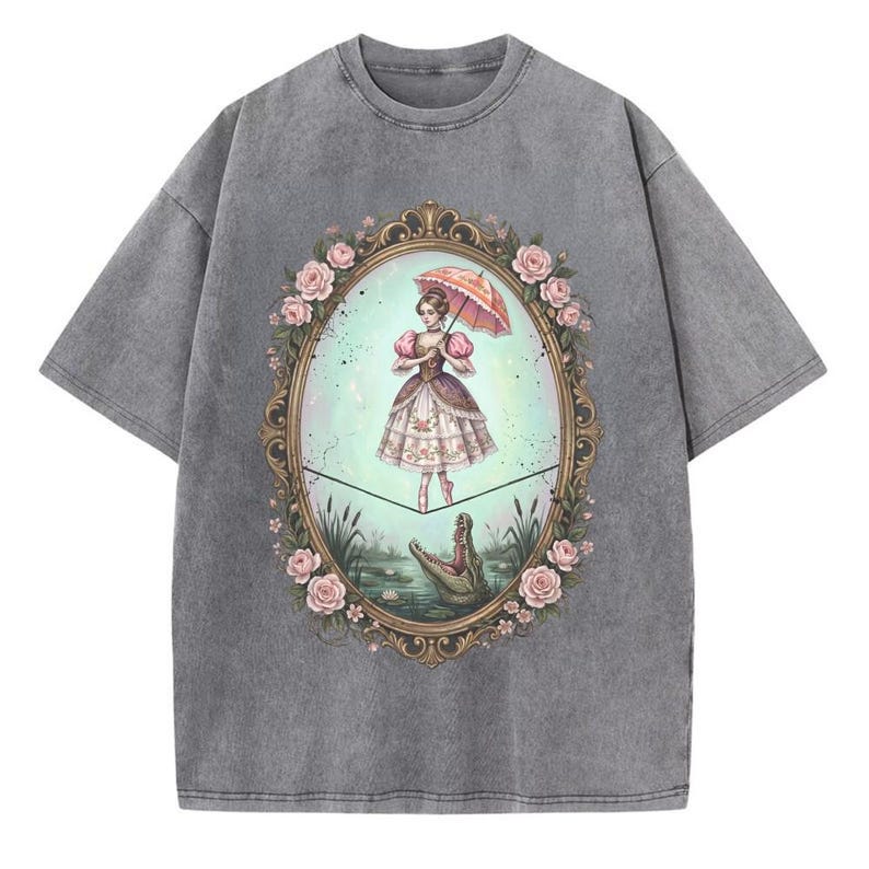May include: Gray t-shirt featuring a framed illustration of a woman with an umbrella, standing on a tightrope above a pond with a crocodile. The frame is adorned with pink roses. The woman is wearing a dress.