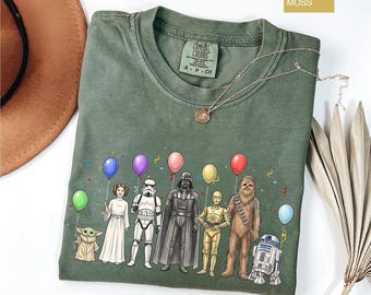 May include: A moss green crew neck t-shirt featuring a graphic print of Star Wars characters including Grogu, Princess Leia, a Stormtrooper, Darth Vader, C-3PO, Chewbacca, and R2-D2 holding colorful balloons and surrounded by confetti.