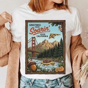 Puede incluir: Camiseta blanca con un dise&ntilde;o gr&aacute;fico de estilo vintage que presenta el texto "Greetings from Soarin' Over California". El dise&ntilde;o incluye el Golden Gate Bridge, un ala delta y una escena de rafting, con personajes de dibujos animados y una bellota.