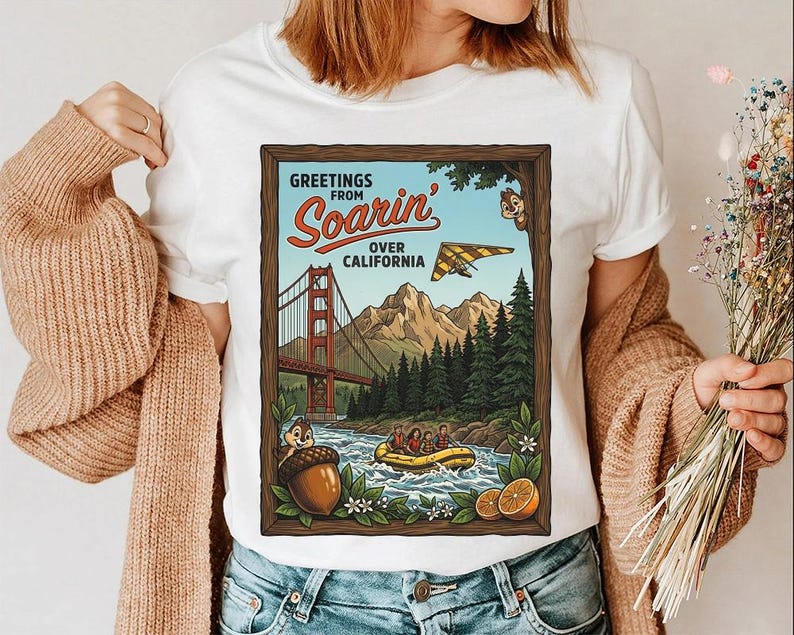 Puede incluir: Camiseta blanca con un dise&ntilde;o gr&aacute;fico de estilo vintage que presenta el texto "Greetings from Soarin' Over California". El dise&ntilde;o incluye el Golden Gate Bridge, un ala delta y una escena de rafting, con personajes de dibujos animados y una bellota.
