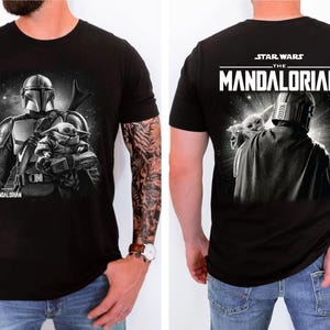 May include: Black t-shirt featuring a graphic of The Mandalorian and Grogu from Star Wars. The front shows the Mandalorian with Grogu, and the back displays the Star Wars: The Mandalorian logo and a similar image. The shirt is short-sleeved.