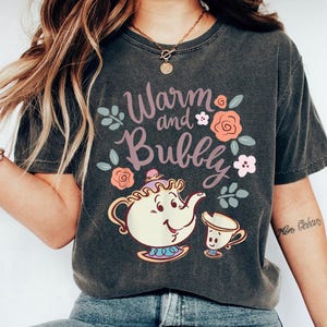 May include: Dark grey t-shirt with a cartoon teapot and teacup design, and the text "Warm and Bubbly". The design includes pink and peach roses and leaves. The shirt has a relaxed fit.