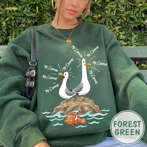 May include: Forest green sweatshirt featuring a cartoon design of two seagulls on a rock with the text "Mr. Connor" and a cartoon orange and white fish. The sweatshirt has a crew neck and long sleeves.