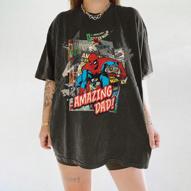 May include: Dark gray t-shirt featuring a comic book style graphic of Spider-Man with the text "AMAZING DAD!". The graphic includes red, blue, and white colors. The shirt has a vintage, worn look.
