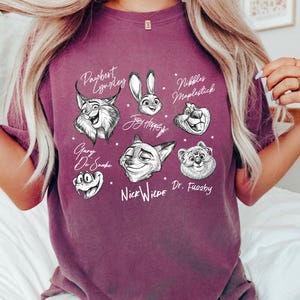 Puede incluir: Una camiseta granate con ilustraciones en blanco y negro de personajes animados. Los nombres de los personajes est&aacute;n escritos encima o debajo de sus retratos. La camiseta es de estilo casual y c&oacute;modo.