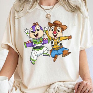 May include: Cream-colored t-shirt featuring a cartoon graphic of two chipmunks. One chipmunk is dressed as Buzz Lightyear, and the other is dressed as a cowboy. The graphic is colorful and detailed, with the chipmunks in dynamic poses.