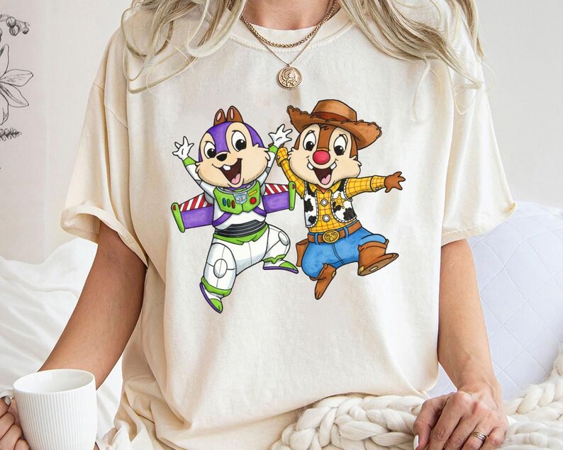 May include: Cream-colored t-shirt featuring a cartoon graphic of two chipmunks. One chipmunk is dressed as Buzz Lightyear, and the other is dressed as a cowboy. The graphic is colorful and detailed, with the chipmunks in dynamic poses.