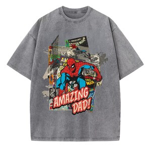 May include: Gray t-shirt featuring a comic book-style graphic of Spider-Man with the text "AMAZING DAD!". The graphic includes red, blue, and white colors. The shirt has short sleeves and a crew neck.