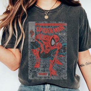 Pode incluir: Uma t-shirt cinzento-carv&atilde;o de manga curta com um gr&aacute;fico de capa de banda desenhada vintage do Spider-Man. A impress&atilde;o mostra a personagem vermelha e azul numa pose agachada com o texto Spider-Man, Torment e The Legend of the Arachknight.