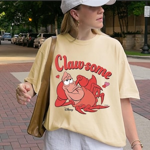 May include: Cream-colored t-shirt featuring a cartoon image of Sebastian the crab from Disney's The Little Mermaid. The shirt has the words "Claw-some" in red script above the crab, with small pink hearts. The Disney logo is at the bottom.