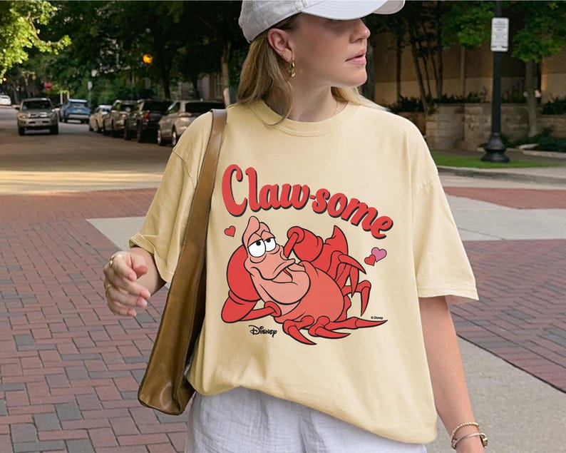 May include: Cream-colored t-shirt featuring a cartoon image of Sebastian the crab from Disney's The Little Mermaid. The shirt has the words "Claw-some" in red script above the crab, with small pink hearts. The Disney logo is at the bottom.