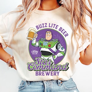 May include: Cream-colored t-shirt featuring a cartoon Buzz Lightyear character holding a beer mug. The design includes the text "Buzz Lite Beer" and "Star Command Brewery" in green and purple, with "Quality Since 1995" text.