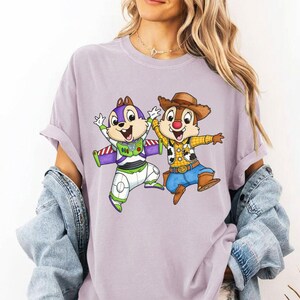 May include: A light purple t-shirt featuring a cartoon graphic of two chipmunks dressed as Buzz Lightyear and Woody. The chipmunks are in a dynamic pose with their arms raised. The shirt has rolled-up sleeves.
