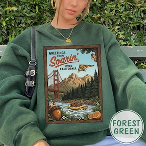 Puede incluir: Sudadera verde bosque con un estampado gr&aacute;fico de estilo vintage. El dise&ntilde;o muestra "Greetings from Soarin' Over California" y representa el Golden Gate Bridge, un ala delta y una escena de rafting. La sudadera tiene cuello redondo.