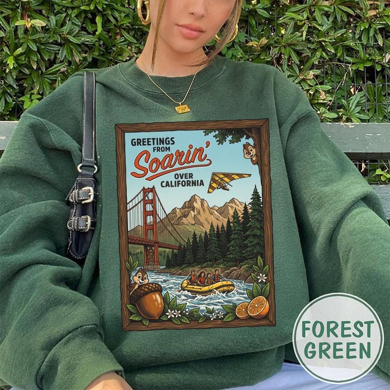 Puede incluir: Sudadera verde bosque con un estampado gr&aacute;fico de estilo vintage. El dise&ntilde;o muestra "Greetings from Soarin' Over California" y representa el Golden Gate Bridge, un ala delta y una escena de rafting. La sudadera tiene cuello redondo.