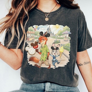 May include: Dark grey t-shirt with a colourful illustration of Nick Wilde and Judy Hopps from Zootopia in a theme park setting. Nick holds a Mickey Mouse-shaped ice cream, and Judy has a soft-serve cone. The background includes a castle and food stalls.