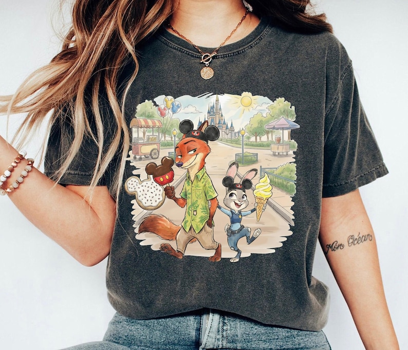 May include: Dark grey t-shirt with a colourful illustration of Nick Wilde and Judy Hopps from Zootopia in a theme park setting. Nick holds a Mickey Mouse-shaped ice cream, and Judy has a soft-serve cone. The background includes a castle and food stalls.