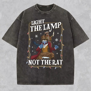 May include: A charcoal grey short-sleeve t-shirt featuring a graphic of a blue character in a top hat and a rat holding a candle. The text reads LIGHT THE LAMP NOT THE RAT, framed by a border decorated with colorful holiday string lights and snowflakes.