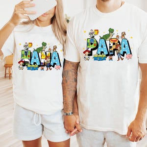 K&ouml;nnte beinhalten: Zwei wei&szlig;e T-Shirts mit den W&ouml;rtern "NANA" und "PAPA" in Blau, verziert mit Zeichentrickfiguren. Die Shirts zeigen einen Dinosaurier, ein Schwein und einen Cowboy. Die Shirts sind wahrscheinlich f&uuml;r eine Familie.
