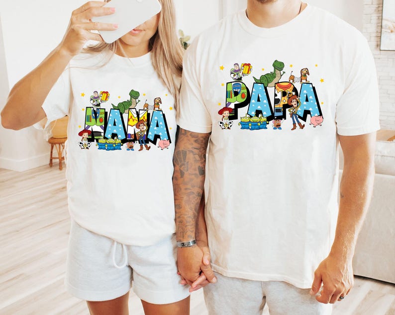 K&ouml;nnte beinhalten: Zwei wei&szlig;e T-Shirts mit den W&ouml;rtern "NANA" und "PAPA" in Blau, verziert mit Zeichentrickfiguren. Die Shirts zeigen einen Dinosaurier, ein Schwein und einen Cowboy. Die Shirts sind wahrscheinlich f&uuml;r eine Familie.