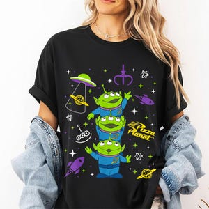May include: Black t-shirt featuring a graphic of three green aliens stacked on top of each other, with the Pizza Planet logo. The design includes rockets, planets, and stars in purple, green, and white.