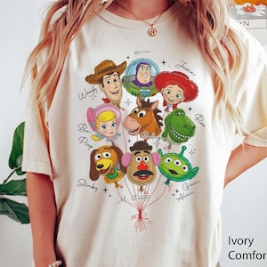 Puede incluir: Camiseta Ivory Comfort Color con un gr&aacute;fico colorido de personajes de Toy Story como globos. Los personajes incluyen a Woody, Buzz Lightyear, Jessie y otros. La camiseta tiene cuello redondo y mangas cortas.