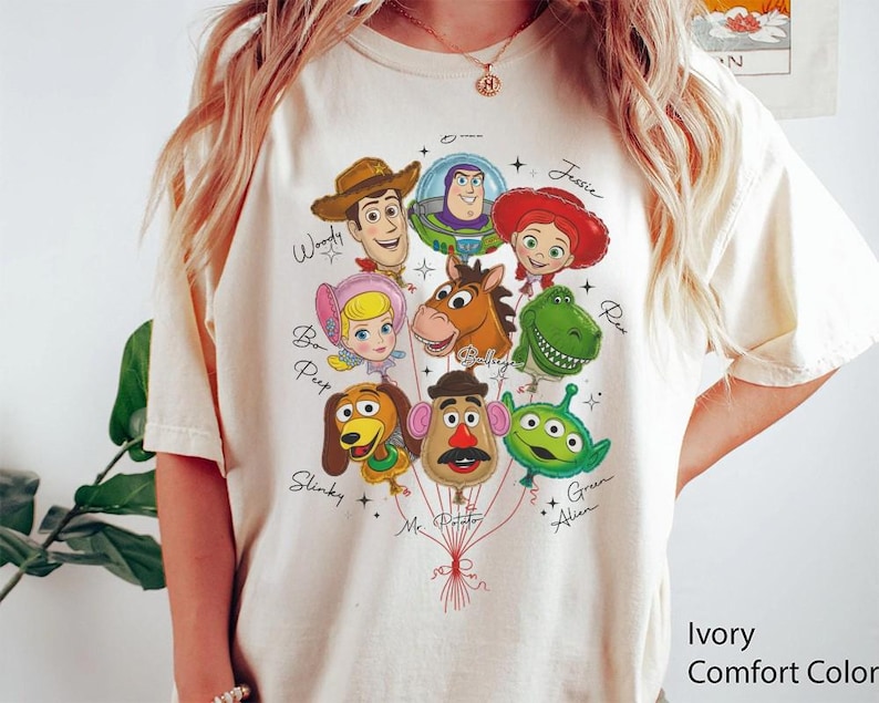 Puede incluir: Camiseta Ivory Comfort Color con un gr&aacute;fico colorido de personajes de Toy Story como globos. Los personajes incluyen a Woody, Buzz Lightyear, Jessie y otros. La camiseta tiene cuello redondo y mangas cortas.