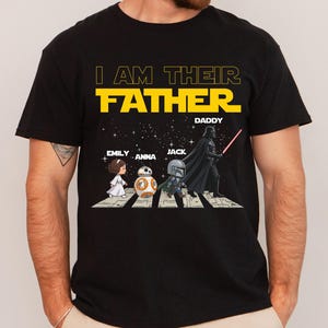 May include: A black short-sleeved t-shirt featuring a graphic of four characters walking on a crosswalk against a starry background. The text reads I AM THEIR FATHER, with names EMILY, ANNA, JACK, and DADDY printed above the illustrated figures.