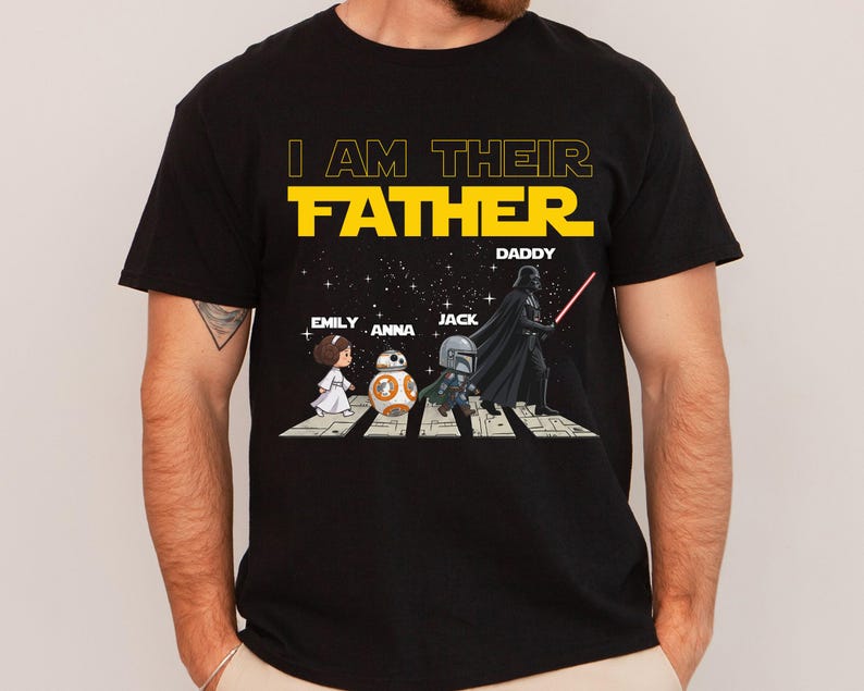 May include: A black short-sleeved t-shirt featuring a graphic of four characters walking on a crosswalk against a starry background. The text reads I AM THEIR FATHER, with names EMILY, ANNA, JACK, and DADDY printed above the illustrated figures.