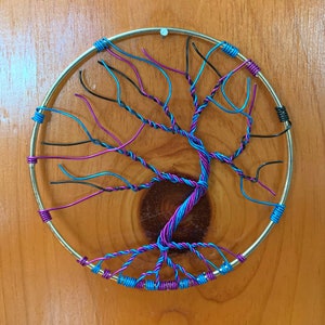 May include: A circular wire art piece depicting a tree with branches in black, teal, and purple wire. The tree is mounted on a wooden background.