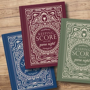 May include: Three hardcover books with ornate covers. The books are in shades of blue, burgundy, and green. Each book features the text "KEEPING SCORE game night" in a circular design, with the names "ABBY & JAMES" at the bottom.