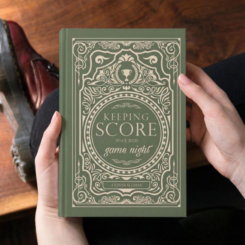 May include: A green book with the words "KEEPING SCORE" and "game night" in a decorative font. The book cover features an ornate design with a trophy and the names "Olivia & Liam".