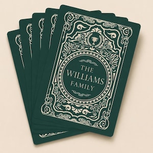 May include: A stack of rectangular playing cards with a dark green background and cream-colored ornate filigree borders. The top card features a central circular frame containing the text THE WILLIAMS FAMILY and a small trophy icon.
