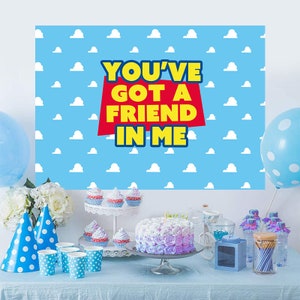May include: A blue and white backdrop with white clouds and the text "You've Got A Friend In Me" in yellow letters on a red rectangle. A table is set with blue and white polka dot paper cups, a two-tiered cake stand with cupcakes, a purple and white frosted cake, and blue and white party favours.