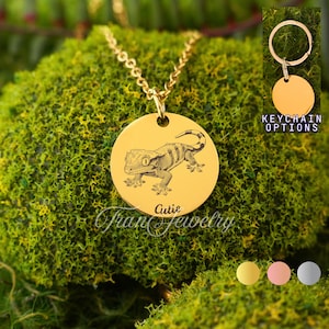 Puede incluir: Collar con colgante dorado con un grabado detallado de un gecko. El colgante es redondo y presenta la palabra "Cutie" y el texto "Fran Jewelry". La imagen también muestra opciones de llavero.