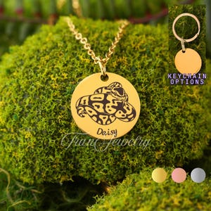 Könnte beinhalten: Eine goldfarbene Halskette mit Anhänger und Kette. Der Anhänger ist eine runde Scheibe mit einer Schlangenillustration und dem eingravierten Namen "Daisy". Die Worte "Fran Jewelry" sind ebenfalls sichtbar. Schlüsselanhänger-Optionen werden angezeigt.