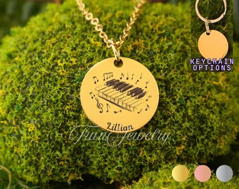 May include: A gold-colored pendant necklace with a piano keyboard design and musical notes. The pendant is round and has the name "Lillian" and "Fran Jewelry" engraved on it. The image also shows keychain options.